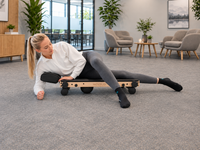 Woman using Movelio Bed Roller Professional for outer hip massage in a bright workplace lounge.