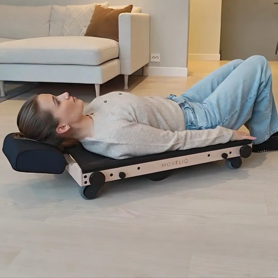 Person lying on the MOVELIO Bed Roller for a deep gravity-based back massage in a modern living room.